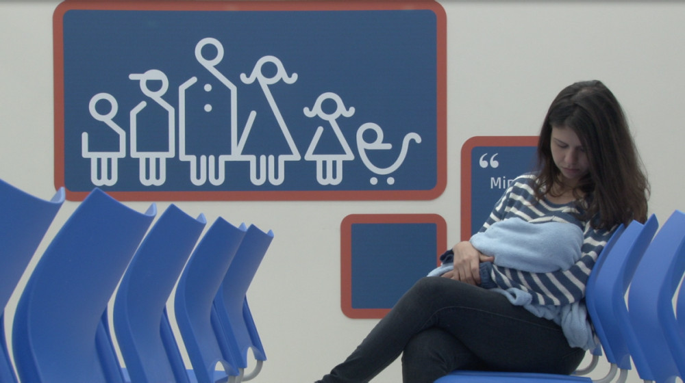 Letícia Bassit segura um bebê sentada entre cadeiras azuis, diante de um painel com figuras de uma família.