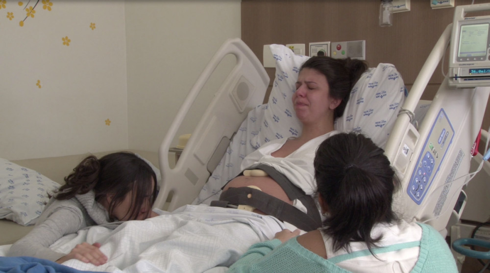 Fotograma de Letícia Bassit em um leito hospitalar, acompanhada por duas pessoas próximas à cama.